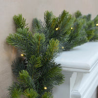 Pre lit Christmas garland with green pine branches and warm white LED lights, draped over a white mantelpiece